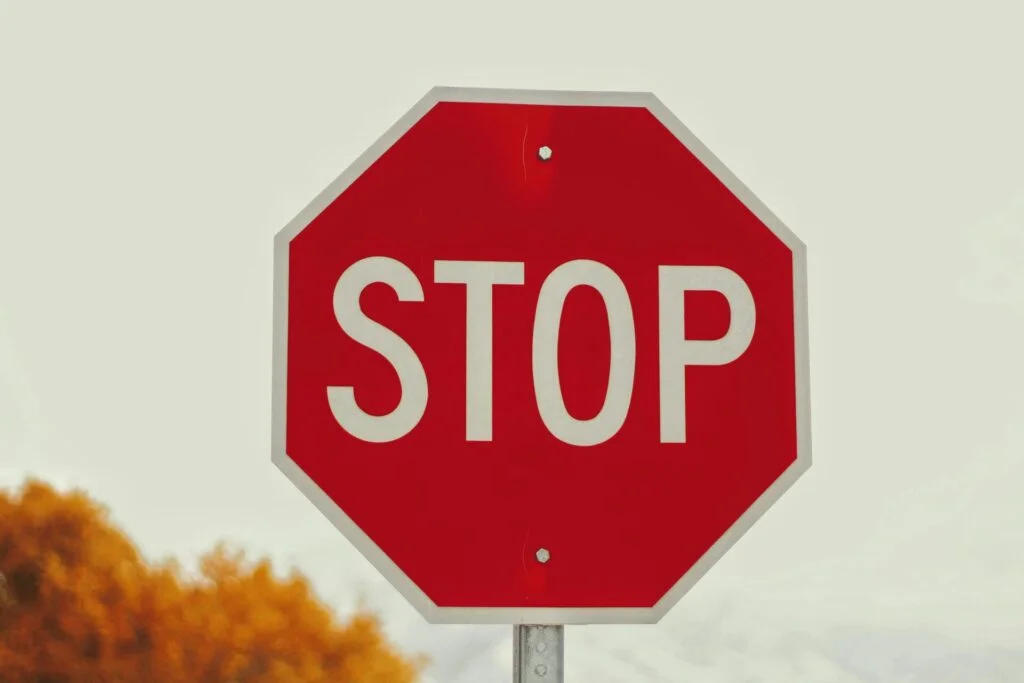 A red octagonal stop sign with the word "STOP" in white letters is pictured against a hazy sky with some orange foliage in the background, reminding viewers of one of the 10 Things You Should Never Do as an Airbnb Host: ignore clear instructions.