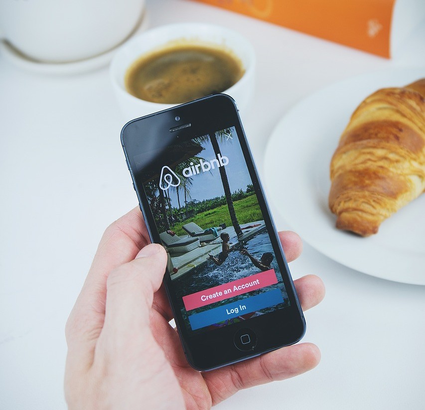 Person holding a smartphone with the Airbnb UK login screen open, next to a coffee and croissant.