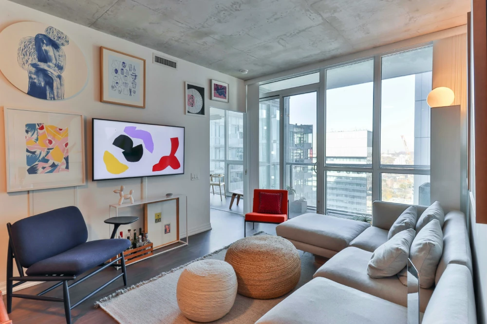 Modern apartment living room with large windows, a grey corner sofa, woven poufs and colourful artwork on the walls